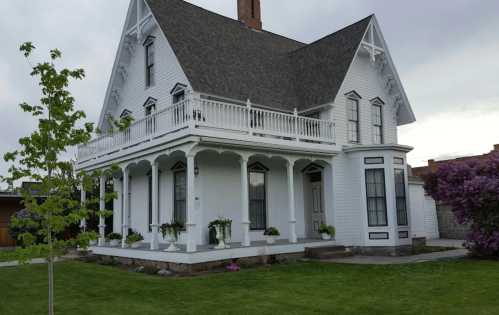 A charming white Victorian-style house with a large porch, surrounded by green grass and blooming flowers.