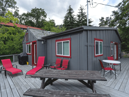 A gray cabin with red accents on a wooden deck, featuring lounge chairs and a picnic table surrounded by greenery.
