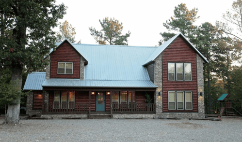 A large, rustic two-story house with a metal roof, stone accents, and a gravel driveway, surrounded by trees.