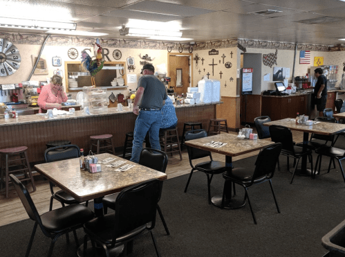 A cozy diner interior with tables, a counter, and staff serving customers. Decor includes various wall items and an American flag.