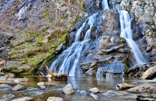 A serene waterfall cascades over rocky cliffs into a calm pool, surrounded by moss and scattered stones.