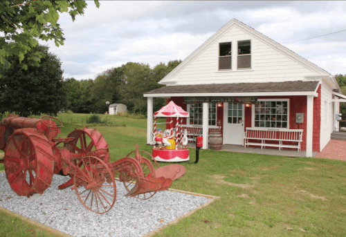 A quaint country store with a red and white exterior, featuring vintage tractors and a colorful carousel in the yard.