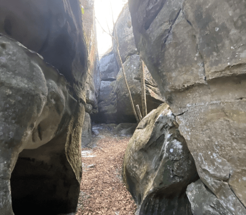 Narrow rocky passageway between large boulders, with sunlight filtering through trees above.