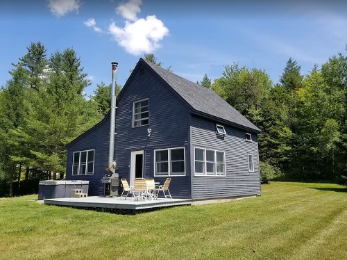 A blue house with a chimney, surrounded by trees and a grassy yard, featuring a deck with outdoor furniture.