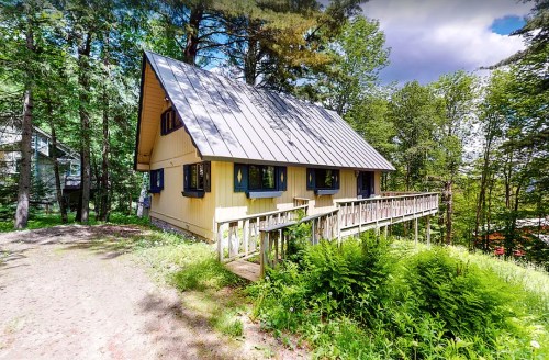 A yellow A-frame house surrounded by trees, with a wooden deck and lush greenery in the foreground.