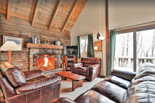Cozy living room with a stone fireplace, leather sofas, and large windows overlooking a wooded area.