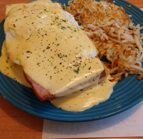 A blue plate with poached eggs topped with creamy sauce and ham, served with crispy hash browns on the side.