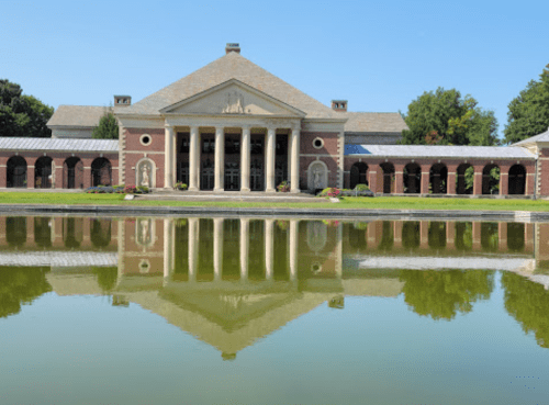 A grand building with columns reflected in a calm pond, surrounded by greenery under a clear blue sky.