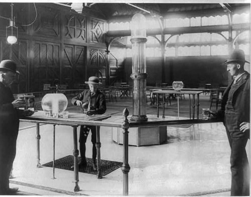 Three men in bowler hats interact in a spacious, vintage room with large glass containers and wooden tables.