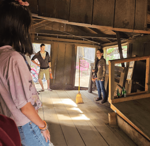 Two people stand in a rustic wooden room, one holding a broom, while another observes from a distance.