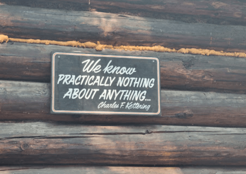 A wooden sign on a log wall reads, "We know practically nothing about anything... Charles F. Kettering."