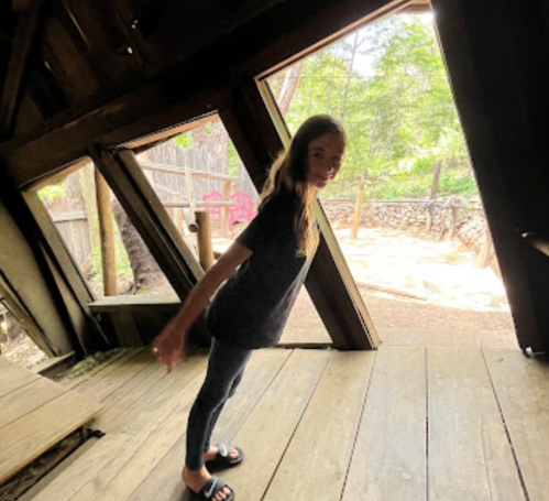 A girl stands inside a wooden structure, leaning slightly with arms outstretched, surrounded by trees and natural light.