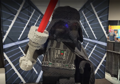 A large LEGO sculpture of a character in a black cape, holding a red lightsaber, set against a geometric background.