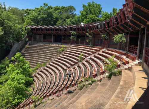 Abandoned outdoor amphitheater with overgrown seating and trees, surrounded by lush greenery under a clear blue sky.