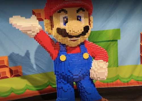 A large LEGO sculpture of Mario, waving, set against a colorful background with blocks and a green pipe.