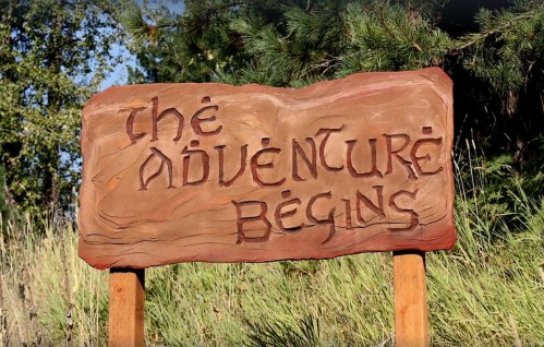 A wooden sign carved with the words "The Adventure Begins," surrounded by greenery.