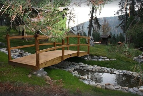 A wooden bridge arches over a small stream, surrounded by lush greenery and mountains in the background.