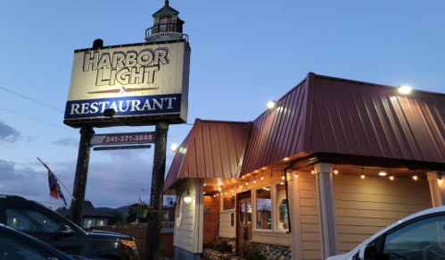 Sign for Harbor Light Restaurant with a lighthouse, illuminated at dusk, featuring a cozy building and parked cars.