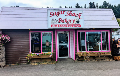 A pink and brown bakery and coffee shop with a sign reading "Sugar Shack Bakery" and open windows adorned with flowers.