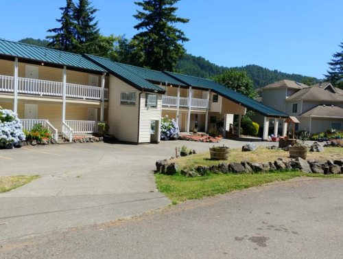 A two-story motel with a green roof, surrounded by trees and flowers, set against a mountainous backdrop.