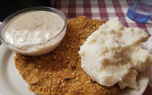 A plate featuring a large fried chicken cutlet, creamy mashed potatoes, and a bowl of gravy on a checkered tablecloth.
