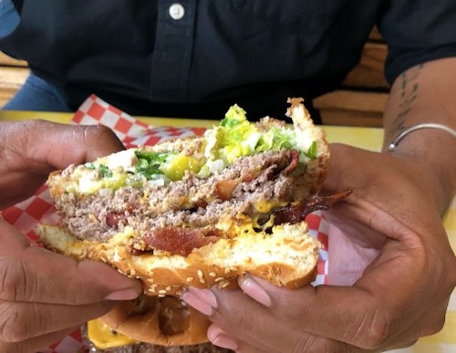 A person holds a large, layered burger with bacon, lettuce, and pickles, showcasing its ingredients.