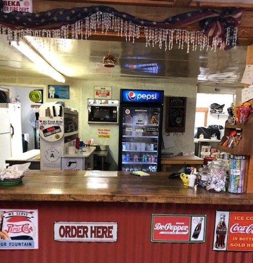 A rustic counter at a diner with a Pepsi fridge, signs, and a menu board, inviting customers to order.
