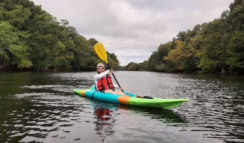 A person in a colorful kayak paddles on a calm river surrounded by lush greenery.