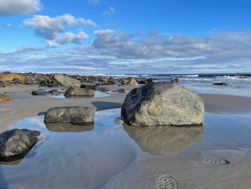 A rocky beach with large stones, reflecting clouds and blue sky, and gentle waves in the background.