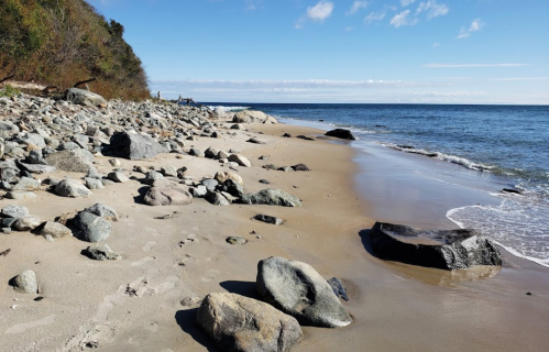 A serene beach scene with smooth rocks, sandy shore, and calm blue water under a clear sky.