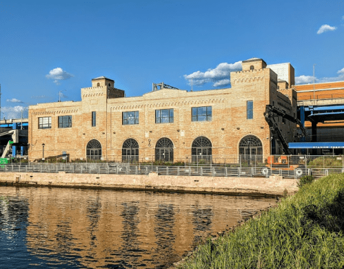 Historic brick building by a river, featuring large windows and a blue sky with scattered clouds.