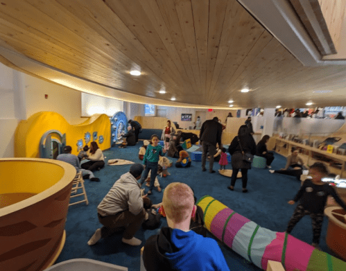 A colorful indoor play area filled with children and adults engaging in various activities on a blue carpet.