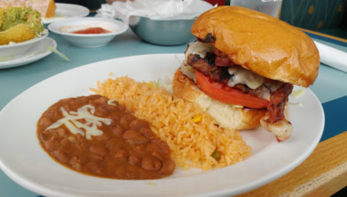 A hearty sandwich with grilled meat, tomato, and cheese, served with rice and beans on a white plate.