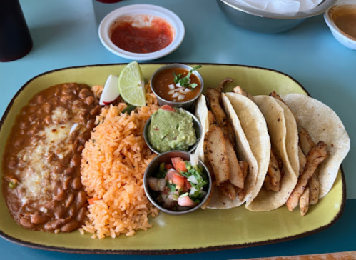 A colorful plate featuring rice, beans, guacamole, salsa, and three chicken tacos with lime wedges.