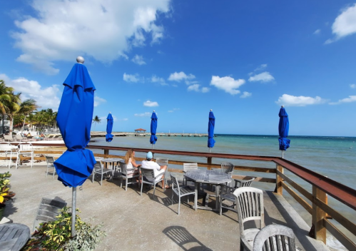 A seaside patio with blue umbrellas, wooden tables, and two people enjoying the view of the ocean and sky.
