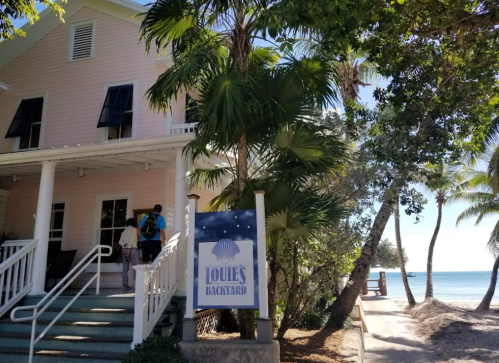 A beachside restaurant with a sign reading "Louie's Backyard," surrounded by palm trees and a view of the ocean.