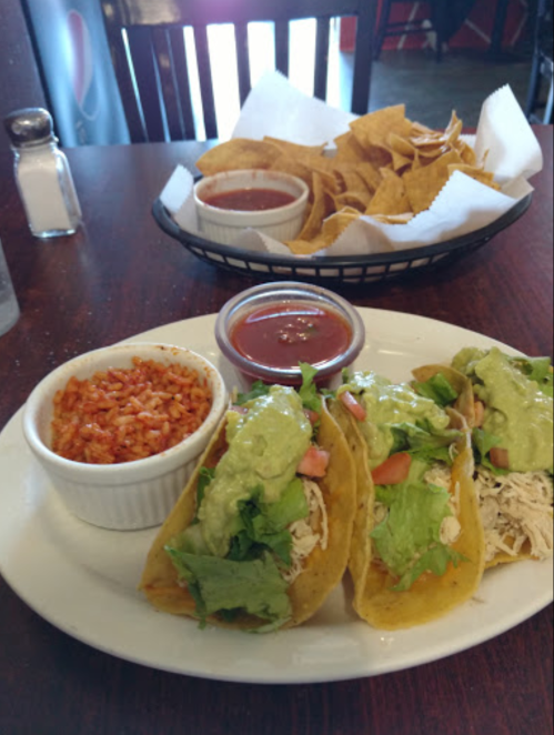 A plate of three tacos with lettuce, tomato, and sauce, served with rice and a side of tortilla chips and salsa.