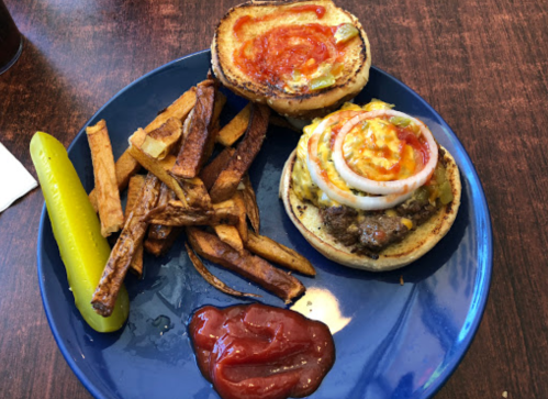 A plate with a burger topped with onions and sauces, served with fries and a pickle on the side.