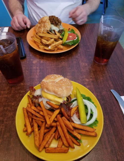 A table with two plates: one with a burger, sweet potato fries, and a pickle; the other with fries and a burger in the background.