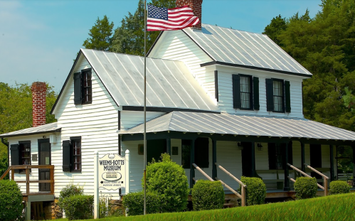 Historic white house with a metal roof, featuring a flag and sign for Weems-Botts Museum, surrounded by greenery.