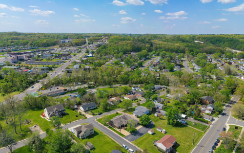 Aerial view of a suburban area with houses, roads, and green trees under a blue sky with scattered clouds.