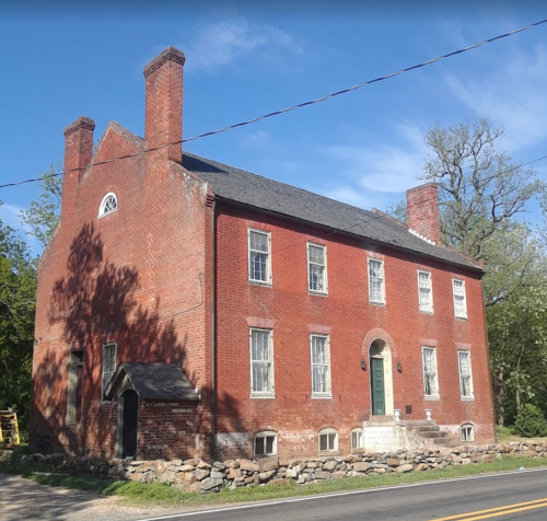 A historic red brick house with multiple windows, two chimneys, and a stone foundation, set against a clear blue sky.