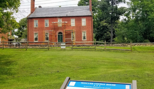 Historic Moncure Conway House under renovation, surrounded by greenery and a wooden fence, with an informational sign in front.