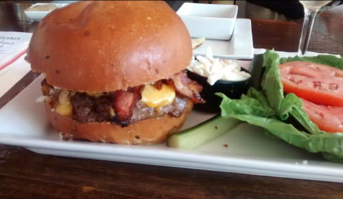 A juicy burger with bacon and cheese, served with lettuce, tomato, pickles, and a side of coleslaw.