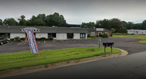 A view of a small antique shop with a colorful sign, surrounded by greenery and a few parked cars.