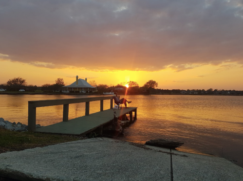 A serene sunset over a calm lake, with a dock in the foreground and a house in the background.