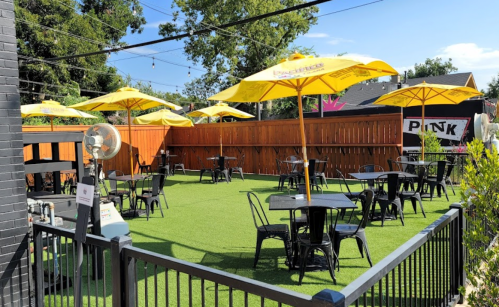 Outdoor patio with black tables and yellow umbrellas, surrounded by a wooden fence and green grass.
