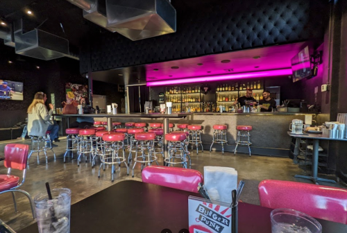 A modern bar with pink lighting, high stools, and a well-stocked shelf behind the counter. Tables in the foreground.