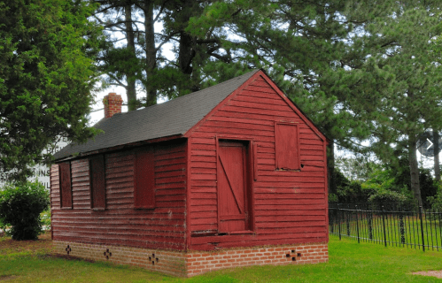A small, red wooden house with boarded windows, surrounded by trees and a fence.