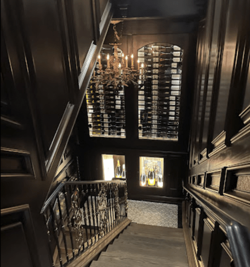 A dark wooden staircase leads down to a wine cellar, featuring a chandelier and shelves filled with wine bottles.